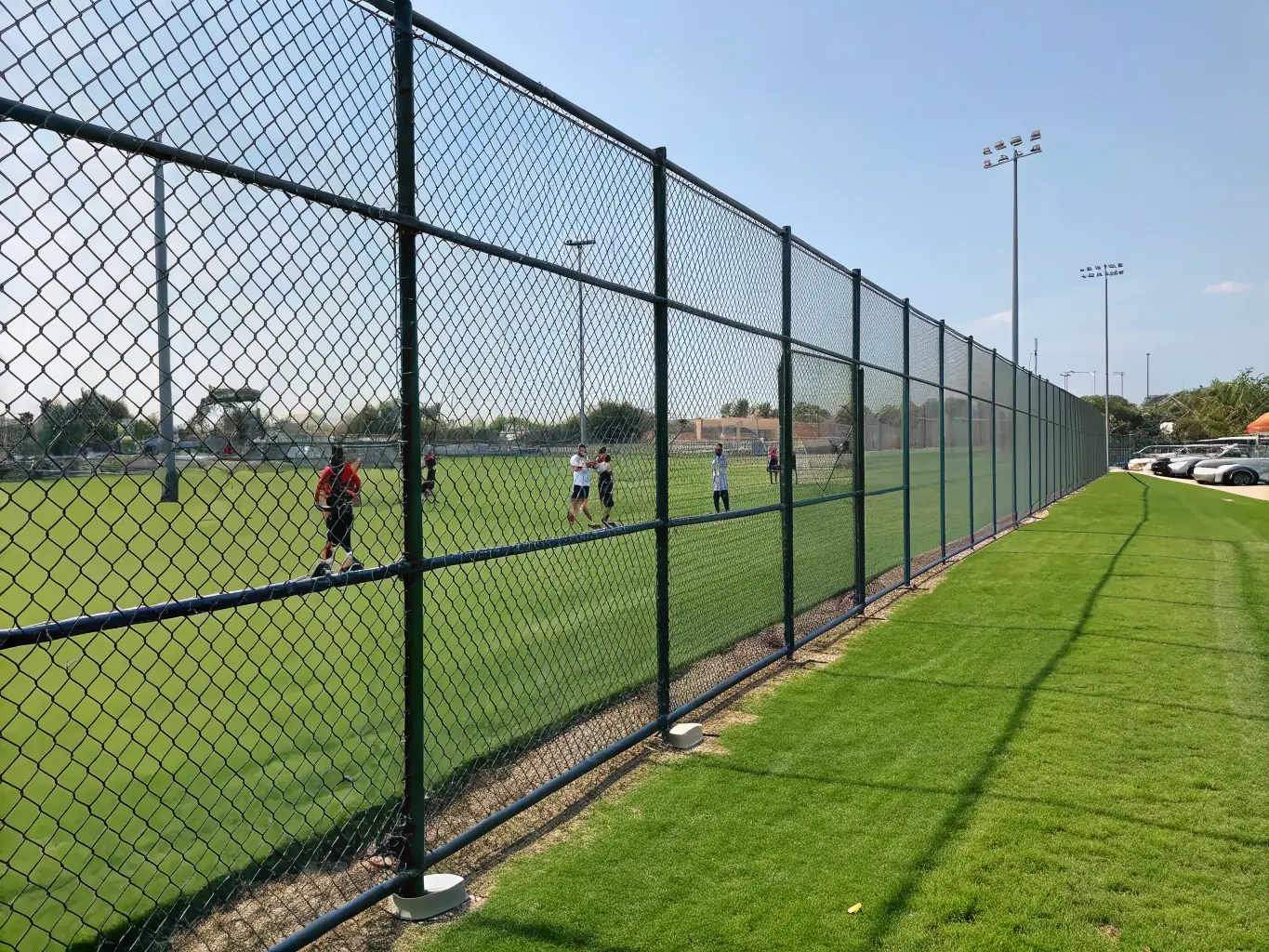 A wide shot of a PVC coated chain link fence enclosing a sports field, demonstrating its versatility and suitability for various applications, with players visible in the background.
